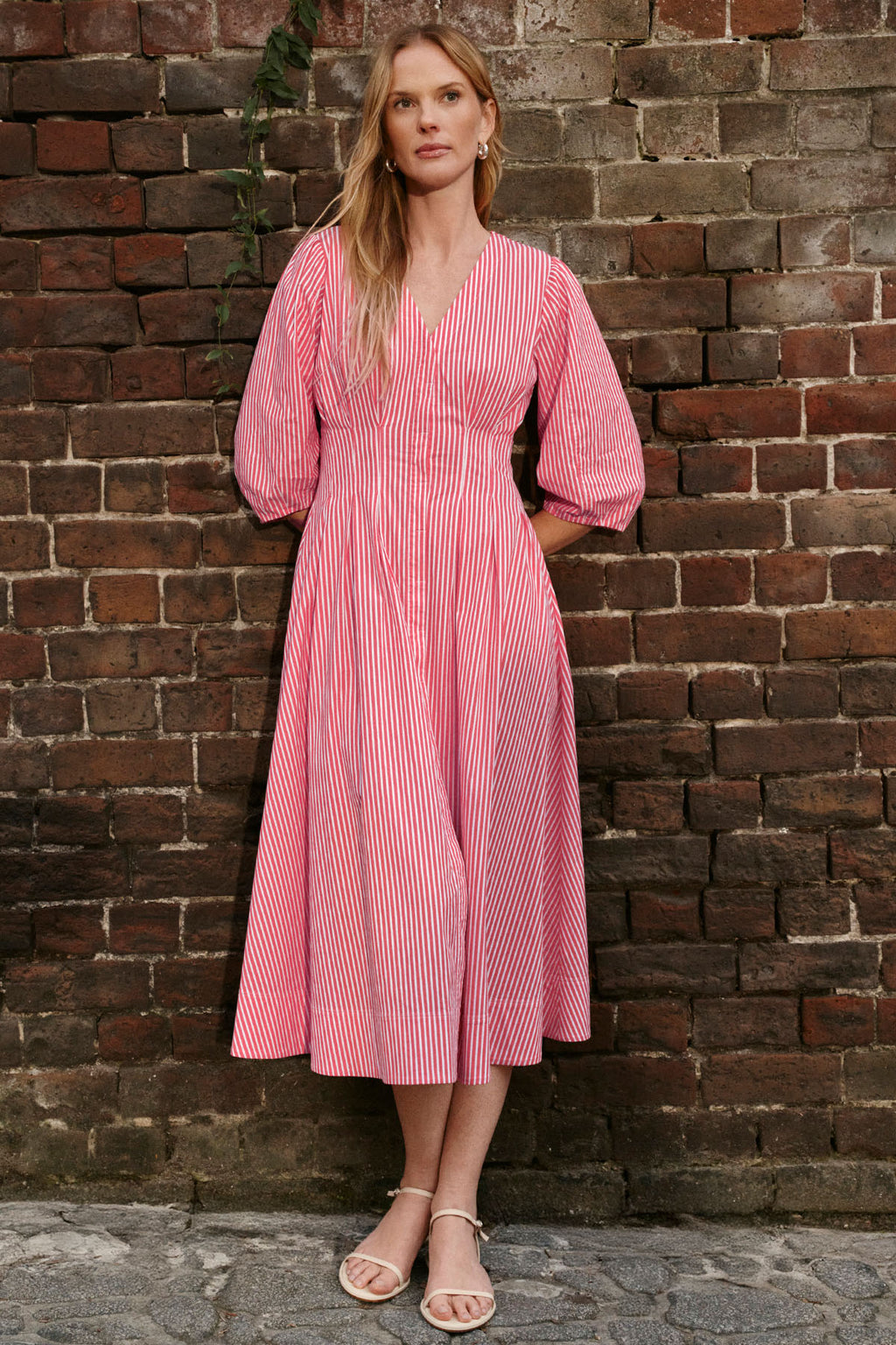 A woman stands against a brick wall, wearing the Sunday Dress in Tart Bengal Stripe—a pink and white striped cotton poplin midi dress with long sleeves, V-neck, and gathered waist—paired with flat sandals.