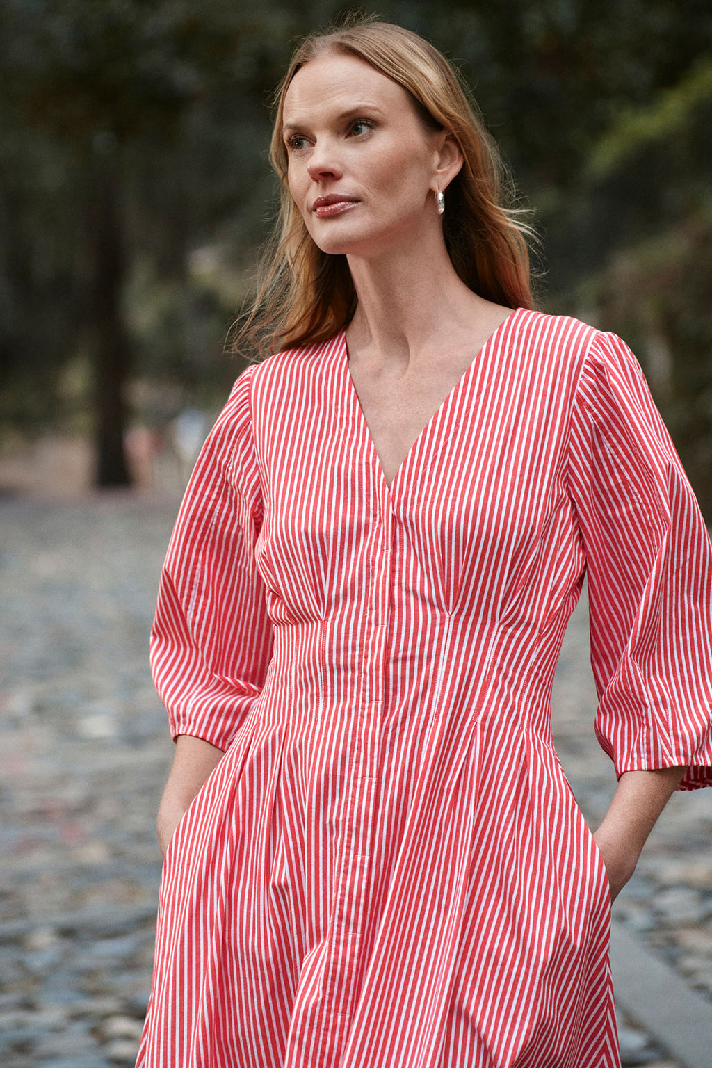 A woman wearing the Sunday Dress in Tart Bengal Stripe stands outdoors on a cobblestone path, gazing calmly to the side.