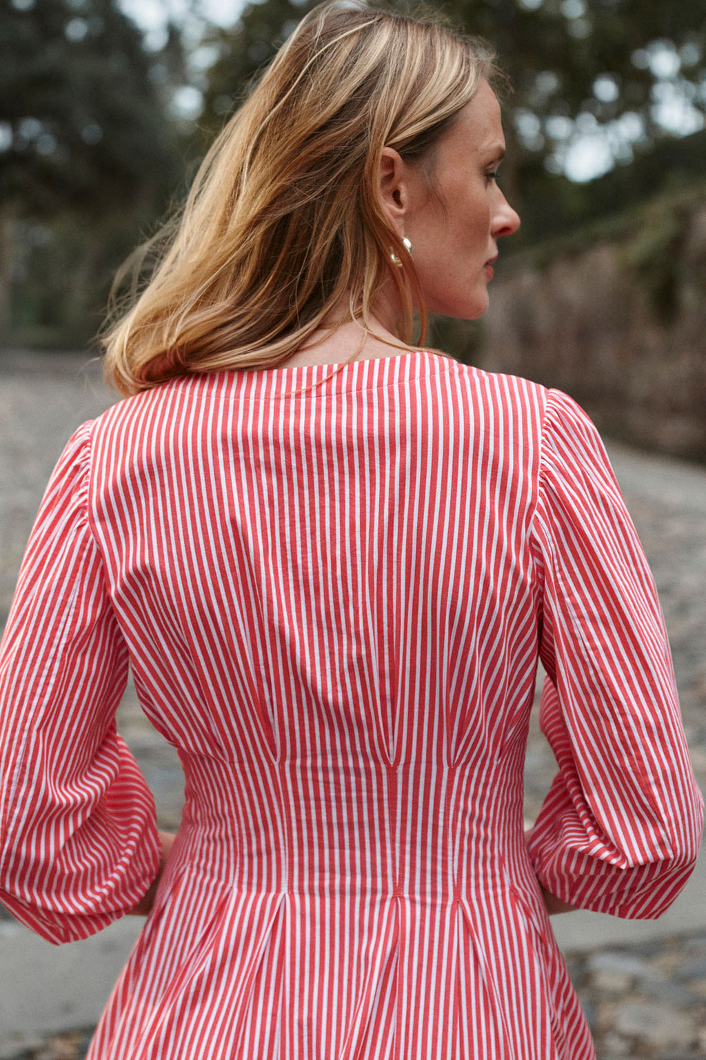 A woman stands outdoors, facing away, wearing the Sunday Dress in Tart Bengal Stripe—a red and white vertically striped cotton poplin dress with long sleeves and a gathered waist. The background features blurred trees and a stone path.
