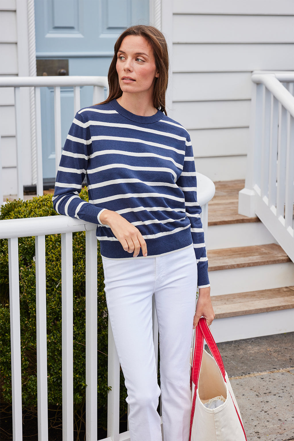 A woman in the Evaline Sweater in Deep Navy Wharf Stripe and white pants stands by a white railing, holding a tote bag, with a light blue door and white house siding behind her.