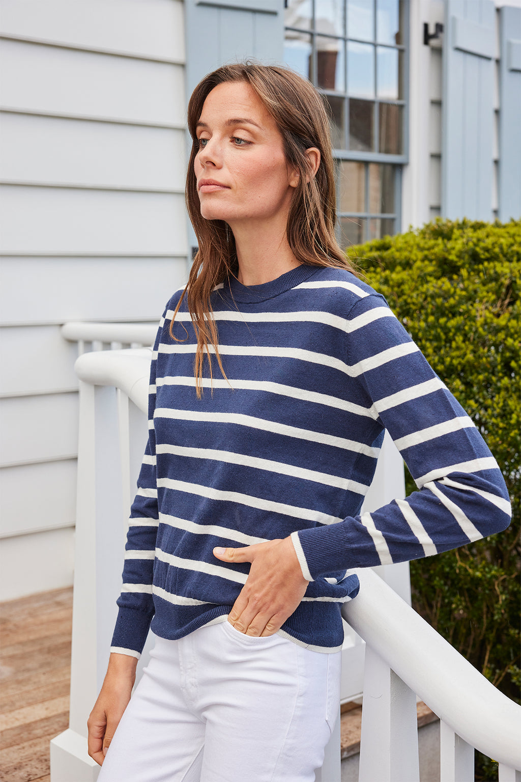 A woman stands outside near white railings, wearing the Evaline Sweater in Deep Navy Wharf Stripe and white pants, with greenery and a house with light blue shutters in the background.