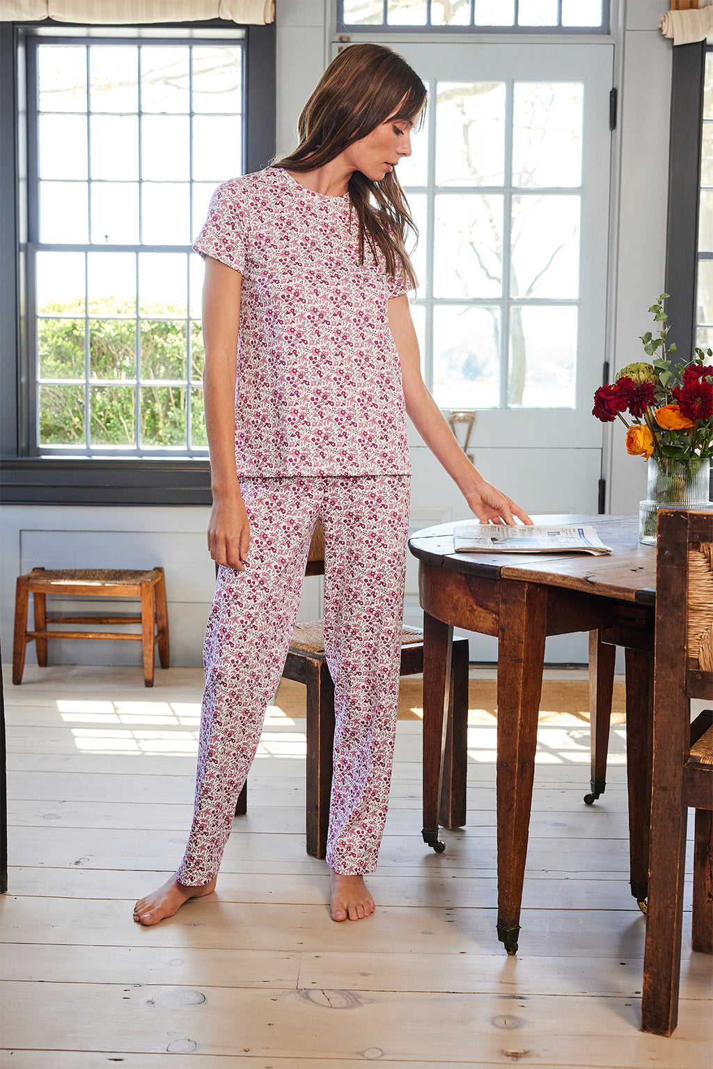 A woman wearing the Pima Crew Bundle in Dahlia Garden Floral stands in a sunlit room, gently touching a wooden table with a vase of flowers and an open book, surrounded by large windows and warm wooden furniture.