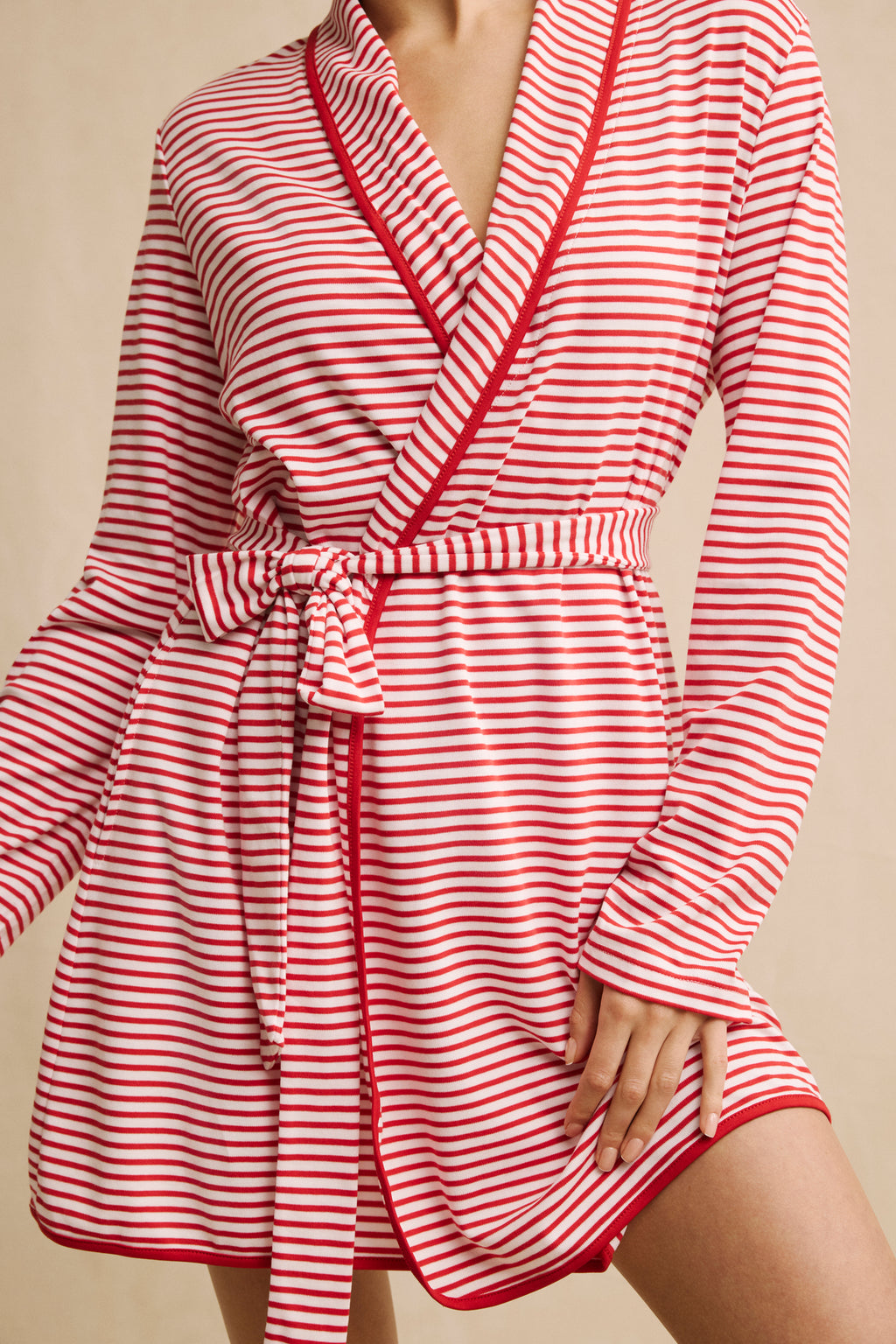 A person models the Pima Short Robe in Classic Red Voyager Stripe, featuring soft Pima cotton, red and white stripes, a belted waist, and a relaxed fit. Their head is out of frame; one hand holds the tie while the other touches their thigh.