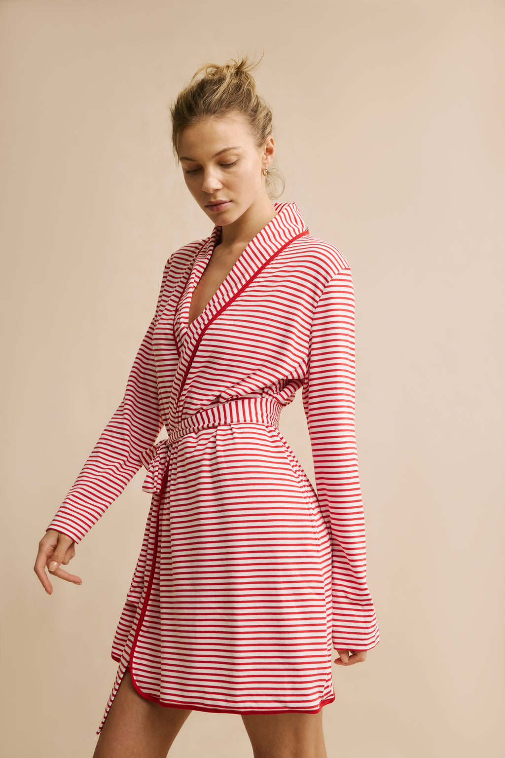 A woman poses against a beige background wearing the Pima Short Robe in Classic Red Voyager Stripe, styled as a wrap dress with long sleeves and a tie belt. Made of soft Pima cotton for luxurious comfort, she looks down with a relaxed expression.