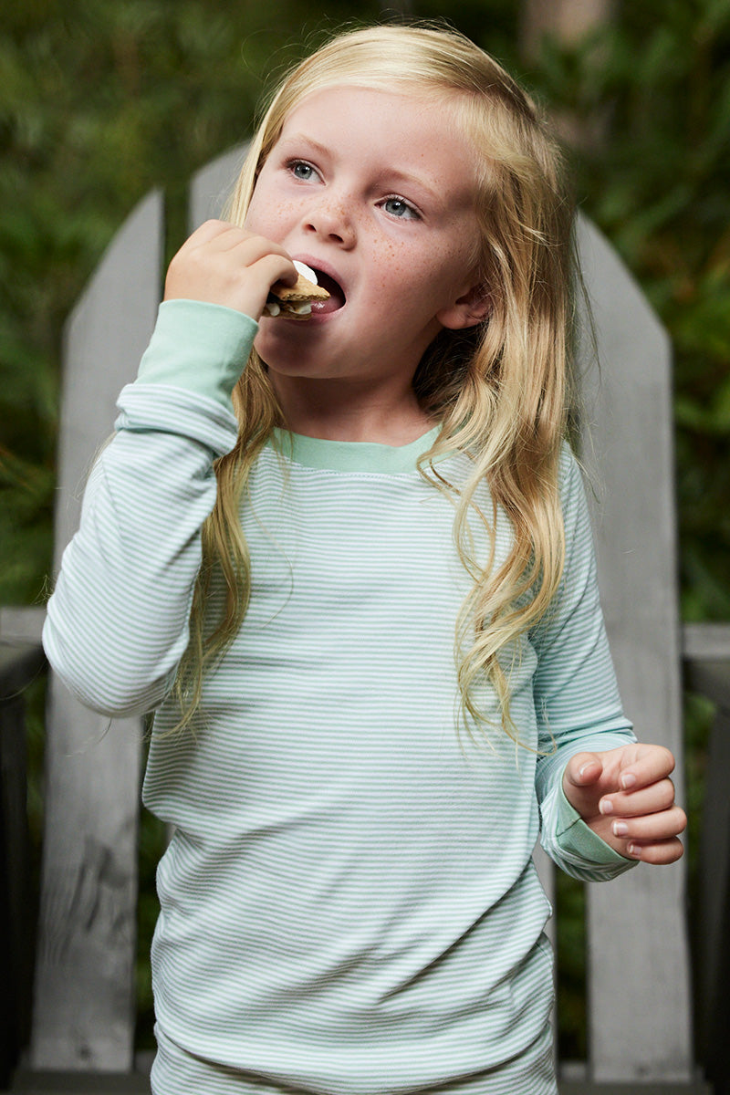 A young child wearing the Kids Long-Long Set in Parisian Green enjoys a marshmallow by a wooden fence with lush greenery, their soft Pima cotton outfit adding charm to this delightful moment.