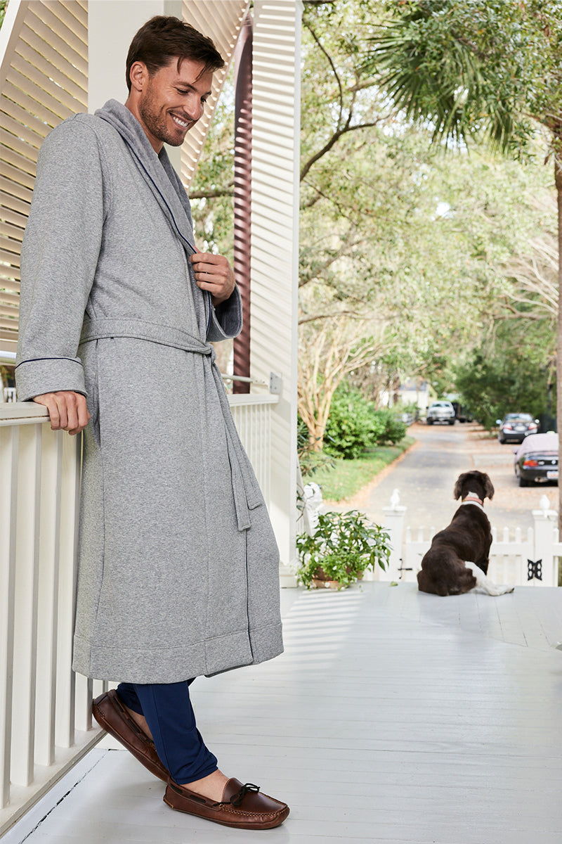 A smiling man in a Mens Cozy Robe in Heather Gray with a turndown collar and fleece lining stands on a porch, leaning against the railing, while a large brown and white dog sits nearby, gazing at the tree-lined street with parked cars.